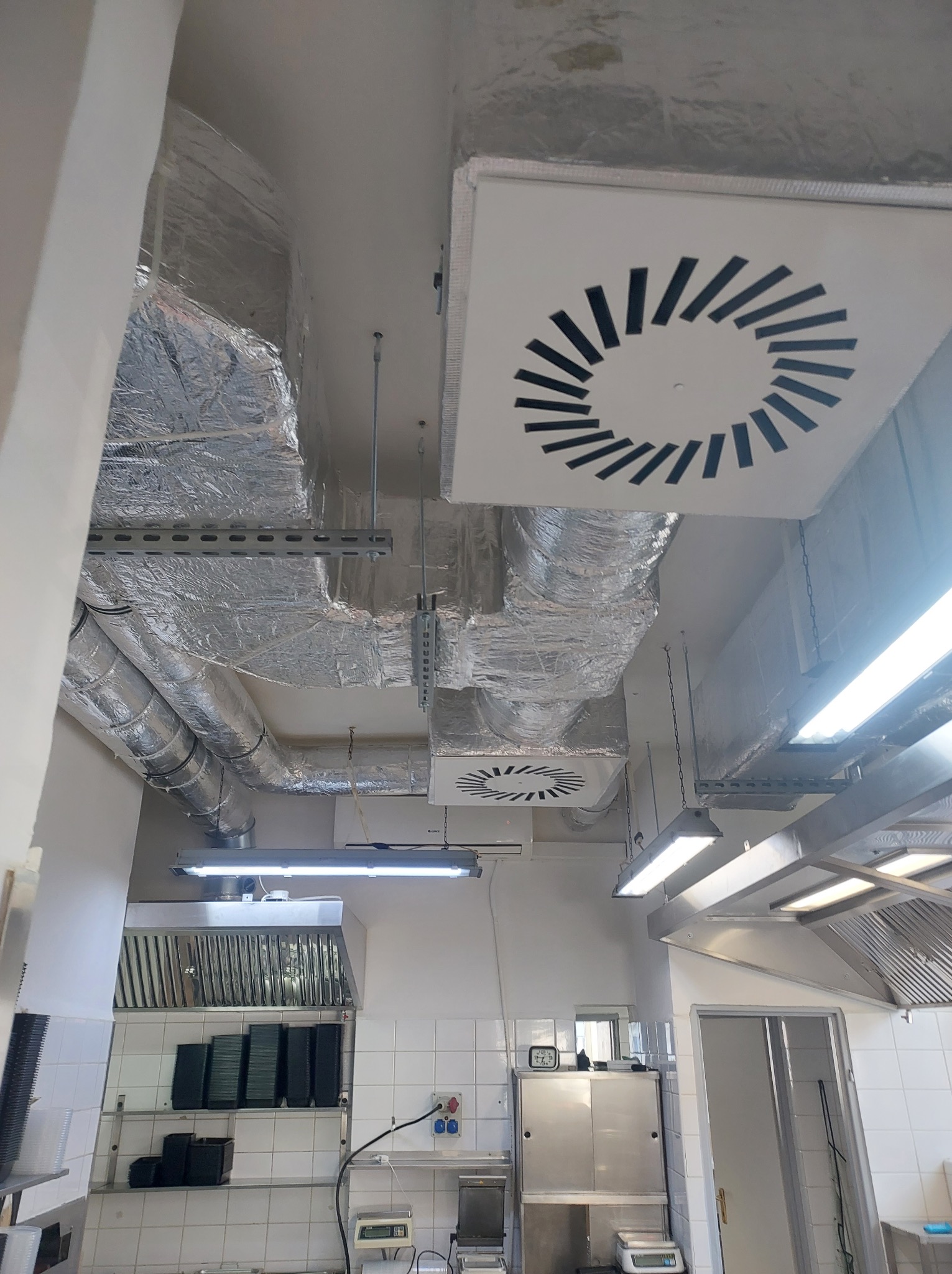 Widok na sufit w kuchni przemysłowej z zamontowanymi kanałami wentylacyjnymi, zabezpieczonymi folią aluminiową oraz kwadratowymi kratkami wentylacyjnymi z charakterystycznym wzorem szczelin...