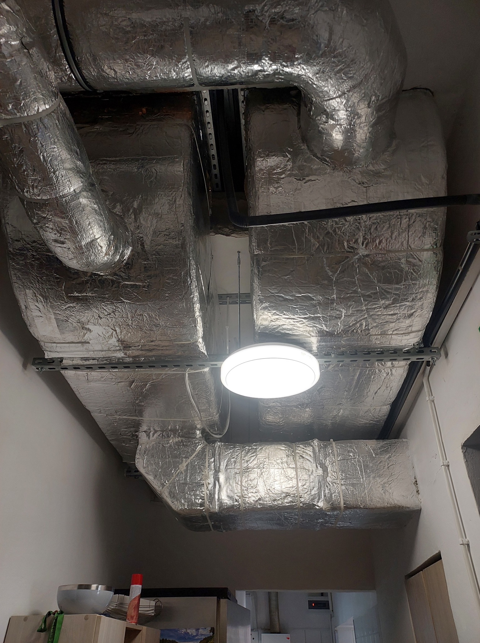 Srebrne, izolowane kanały wentylacyjne pod sufitem, zamontowane na metalowych szynach, oświetlone okrągłą lampą sufitową, widok z dołu w pomieszczeniu.