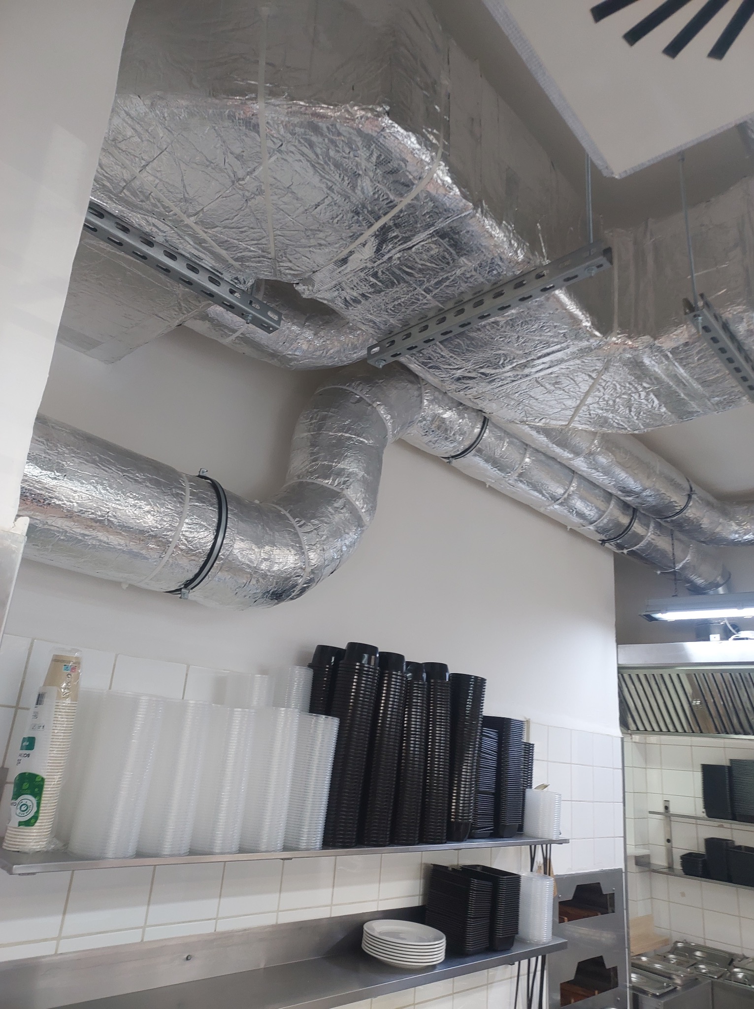 Izolowane kanały wentylacyjne zamontowane pod sufitem w lokalu gastronomicznym, widoczne rury z srebrną folią, metalowe wsporniki i półka z pojemnikami.