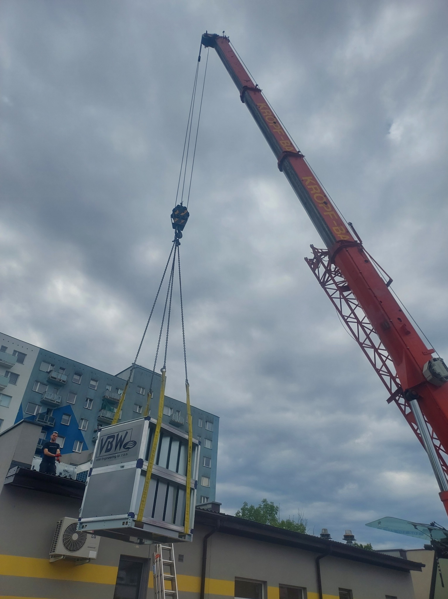 Montaż centrali wentylacyjnej na dachu budynku za pomocą dźwigu KROPF-BA, pracownik nadzoruje proces, widoczna klimatyzacja zewnętrzna LG na ścianie budynku.