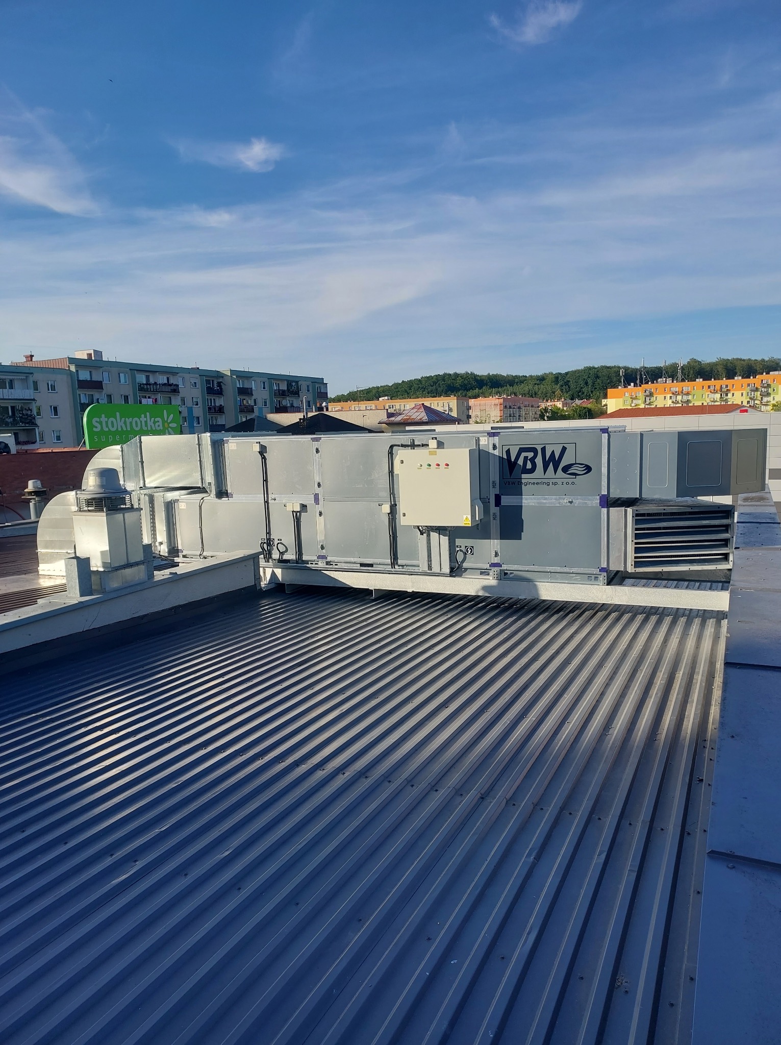 Szara centrala wentylacyjna z logo VBW Engineering zamontowana na dachu z blachy trapezowej; w tle widoczne budynki miejskie i błękitne niebo.