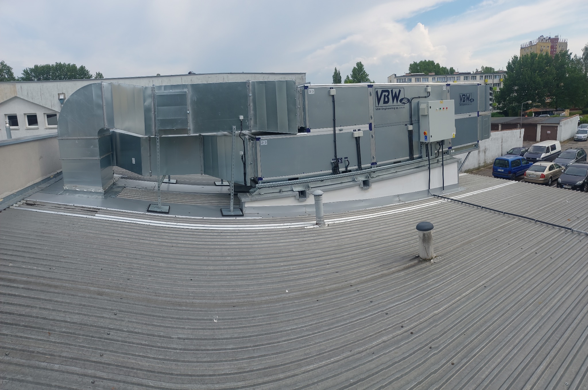Rozległy system kanałów wentylacyjnych z logo firmy na dachu budynku, widok z góry na falisty blaszany dach i zaparkowane samochody w tle.