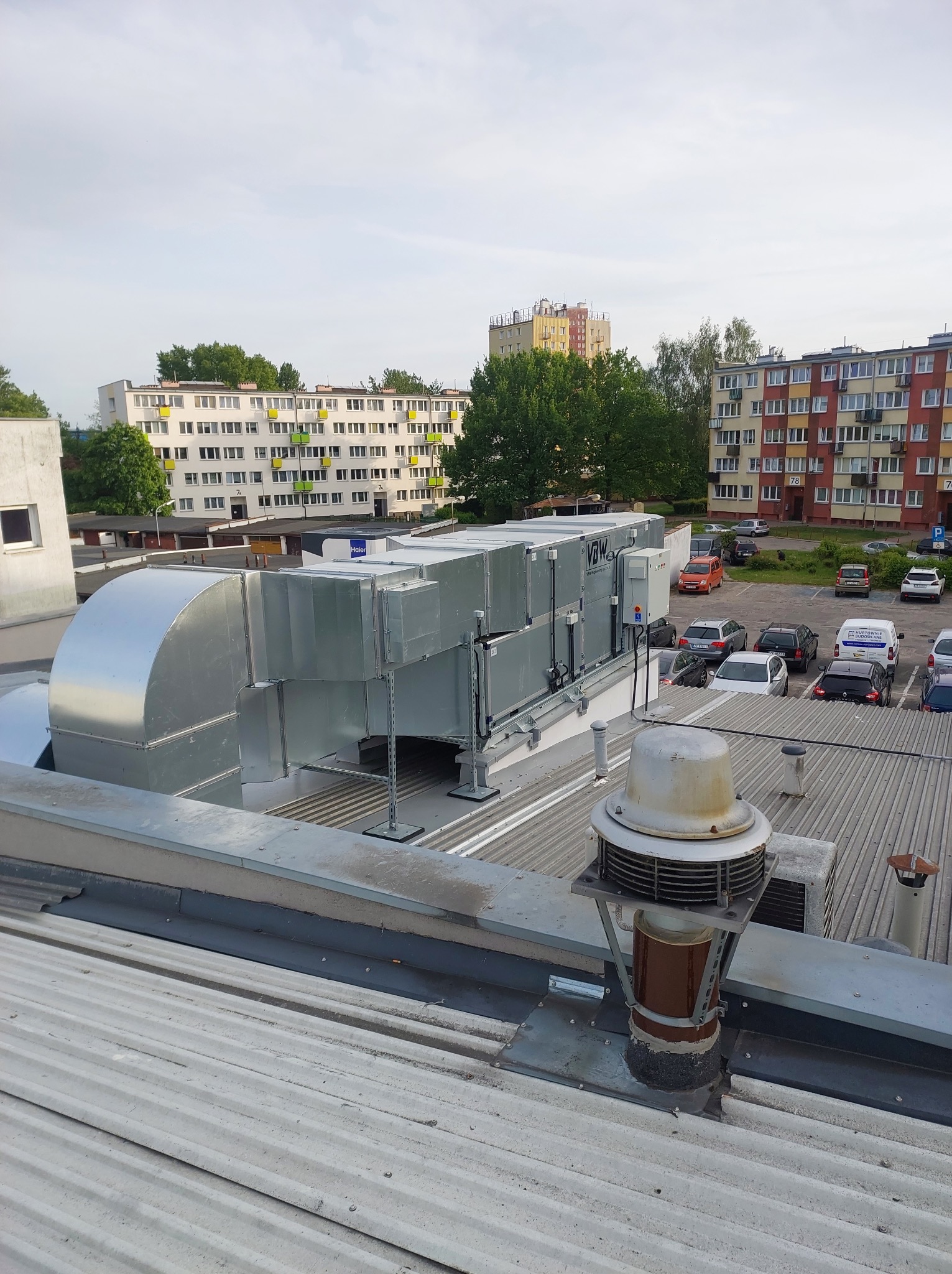 System wentylacyjny na dachu budynku z widokiem na parking i bloki mieszkalne w tle.