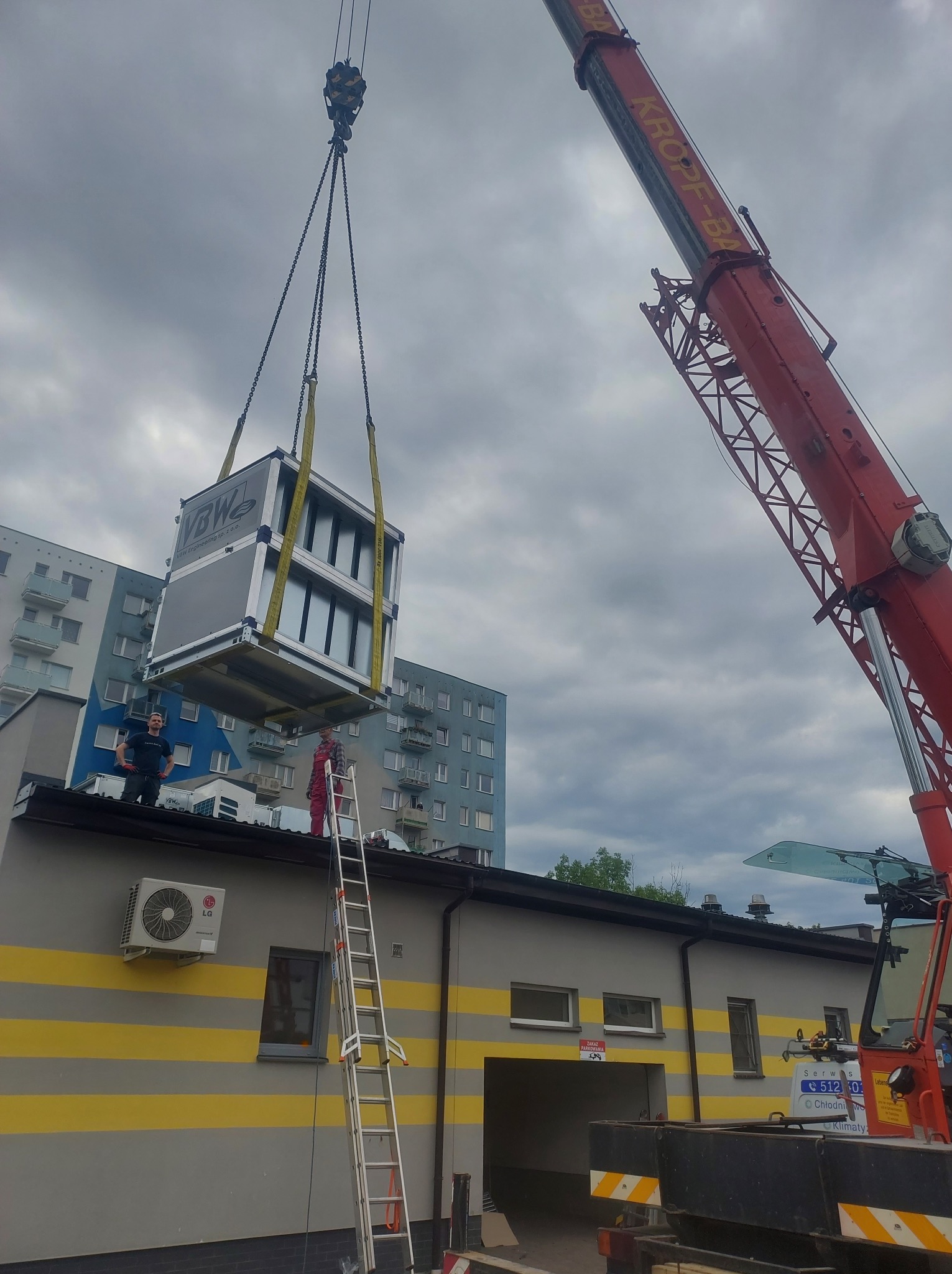 Montaż centrali wentylacyjnej na dachu budynku przy użyciu dźwigu Kropf-BA, widoczni dwaj pracownicy na dachu i drabina oparta o budynek.