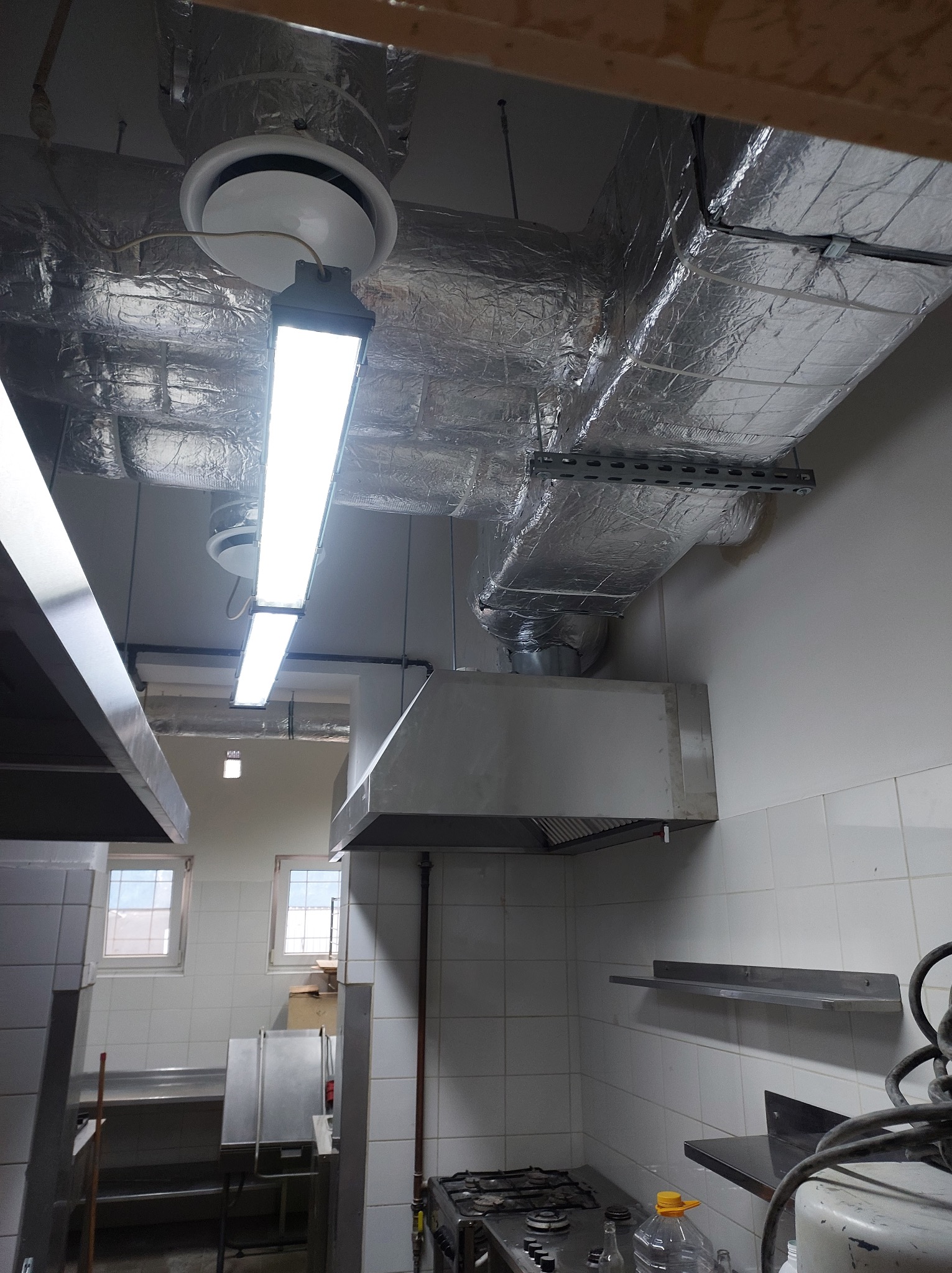 Widok na sufit w kuchni przemysłowej z zainstalowaną wentylacją, w tym kanały owinięte folią aluminiową, okrągłe nawiewniki i lampy jarzeniowe.