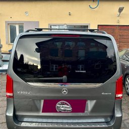 Detailing Olivier Marciniak - Szary Mercedes V300d 4MATIC z widocznym logotypem i naklejką z grafiką samochodu, zaparkowany przed żółtym budynkiem z widocznym odbiciem budynku w szybie.