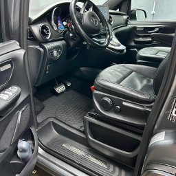 Detailing Olivier Marciniak - Wnętrze samochodu Mercedes-Benz V-klasy, widok od strony otwartych drzwi kierowcy, czarna skórzana tapicerka fotela z elektryczną regulacją, aluminiowe nakładki na pedały, gumowe dywaniki z wzorem...