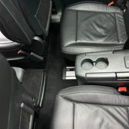 Wnętrze samochodu z czarną skórzaną tapicerką, widoczne fotele z siatkowymi kieszeniami, podłokietnik z uchwytami na napoje i czerwone pasy bezpieczeństwa.