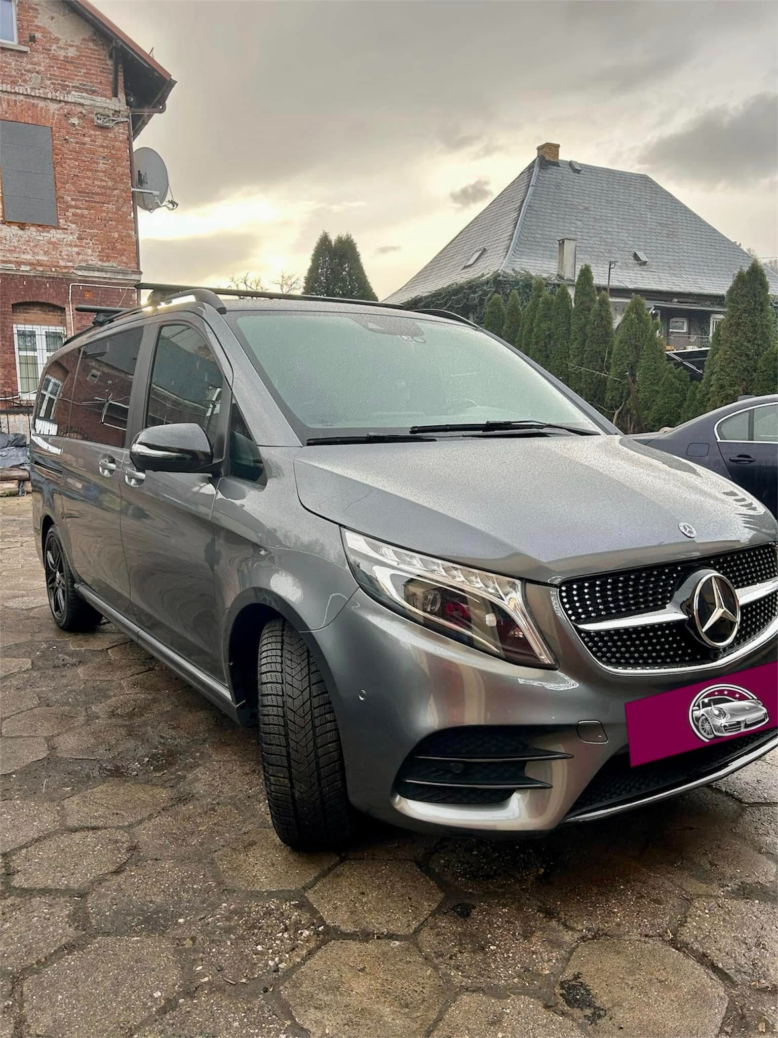 Szary Mercedes-Benz V-klasa zaparkowany na brukowanej nawierzchni przed budynkiem z cegły w pochmurny dzień.