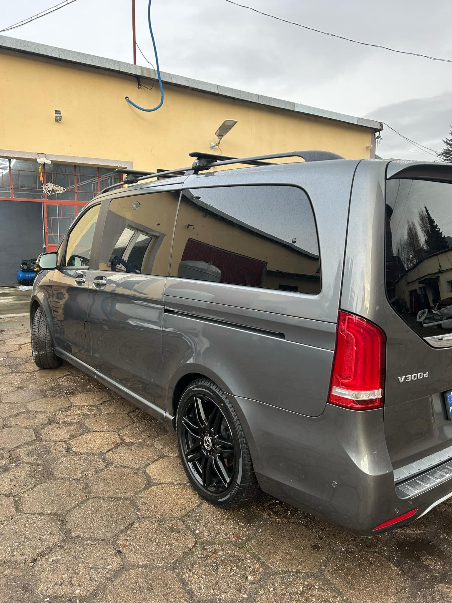 Szary Mercedes V300d z relingami dachowymi po myciu, mokry lakier i czarne felgi na tle żółtej ściany myjni.