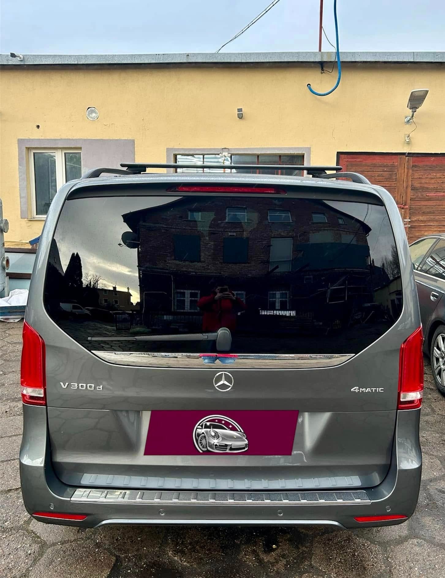 Szary Mercedes V300d 4MATIC z widocznym logotypem i naklejką z grafiką samochodu, zaparkowany przed żółtym budynkiem z widocznym odbiciem budynku w szybie.