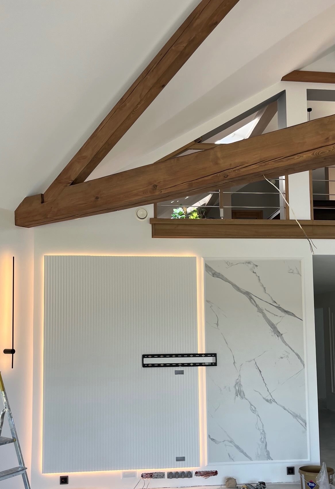 Wnętrze z drewnianymi belkami stropowymi, podświetlaną ścianą z pionowymi żłobieniami, panelem imitującym marmur, uchwytem do telewizora i widocznymi przewodami elektrycznymi w trakcie prac...