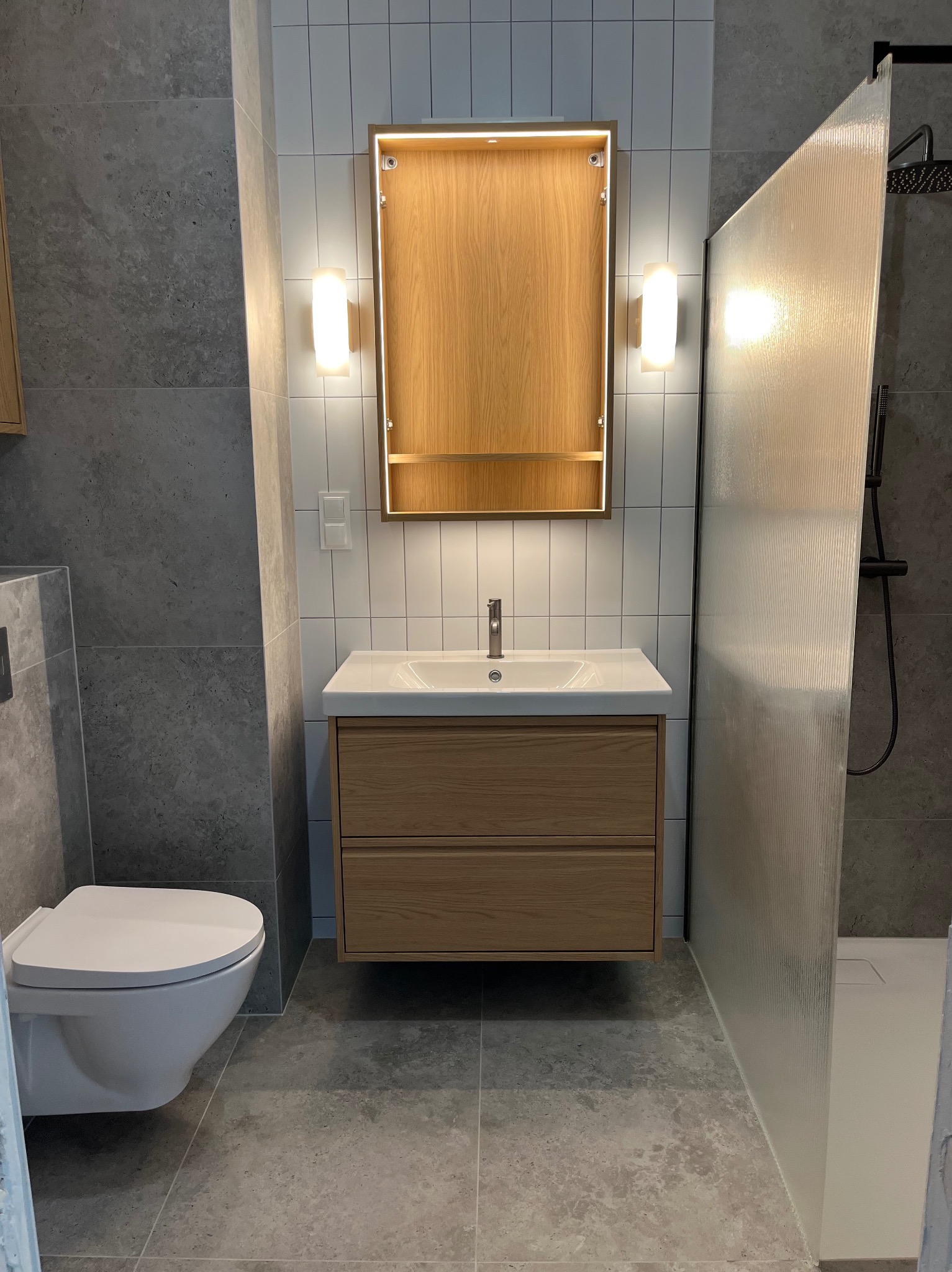 Nowoczesna łazienka z wiszącą szafką pod umywalką z drewnianymi frontami, podświetlanym lustrem w drewnianej ramie, toaletą i kabiną prysznicową z ryflowaną szybą.