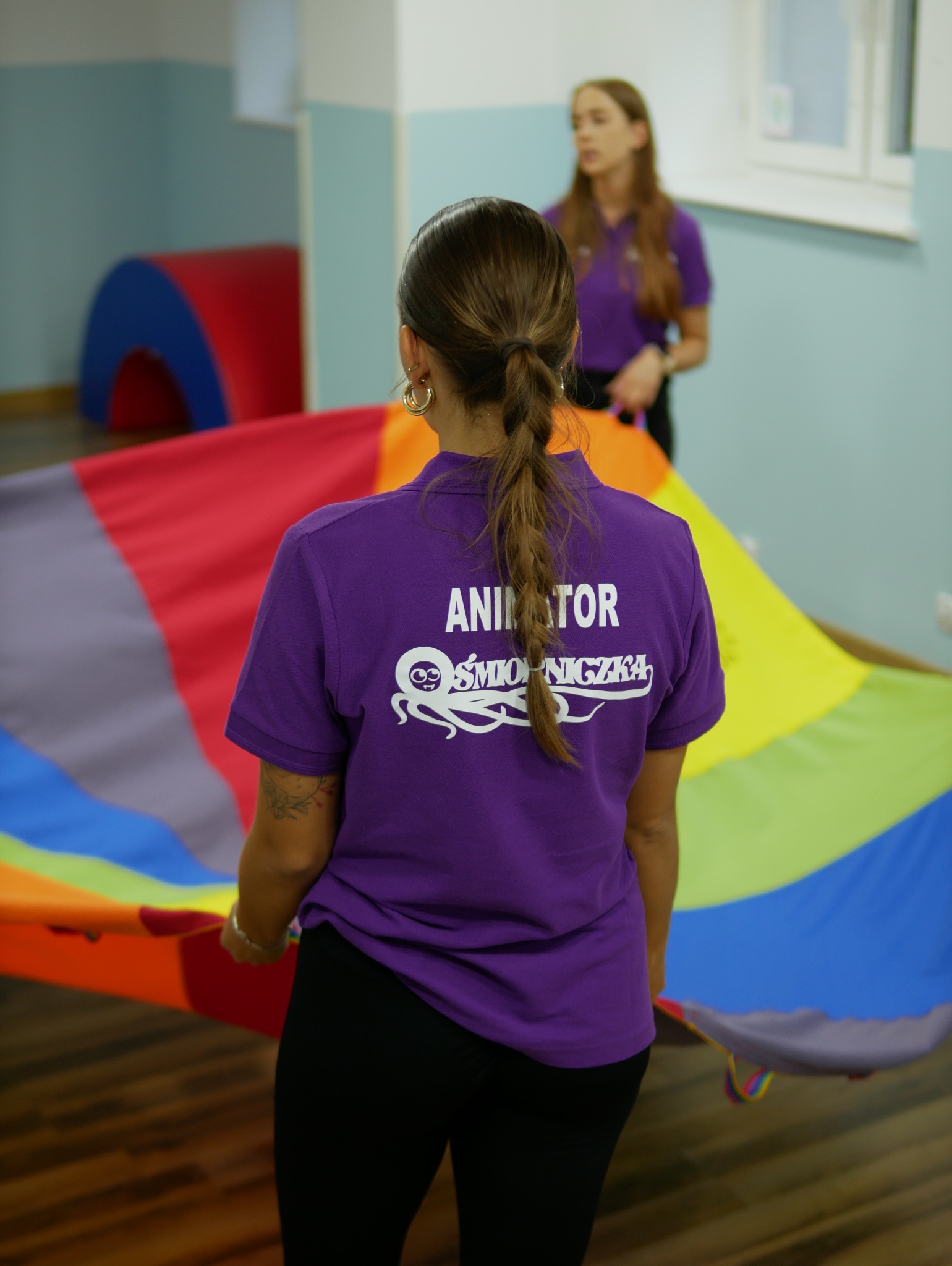 Kobieta w fioletowej koszulce z napisem 'Animator' i logo firmy trzyma kolorową chustę animacyjną, w tle druga animatorka i elementy wyposażenia sali zabaw.