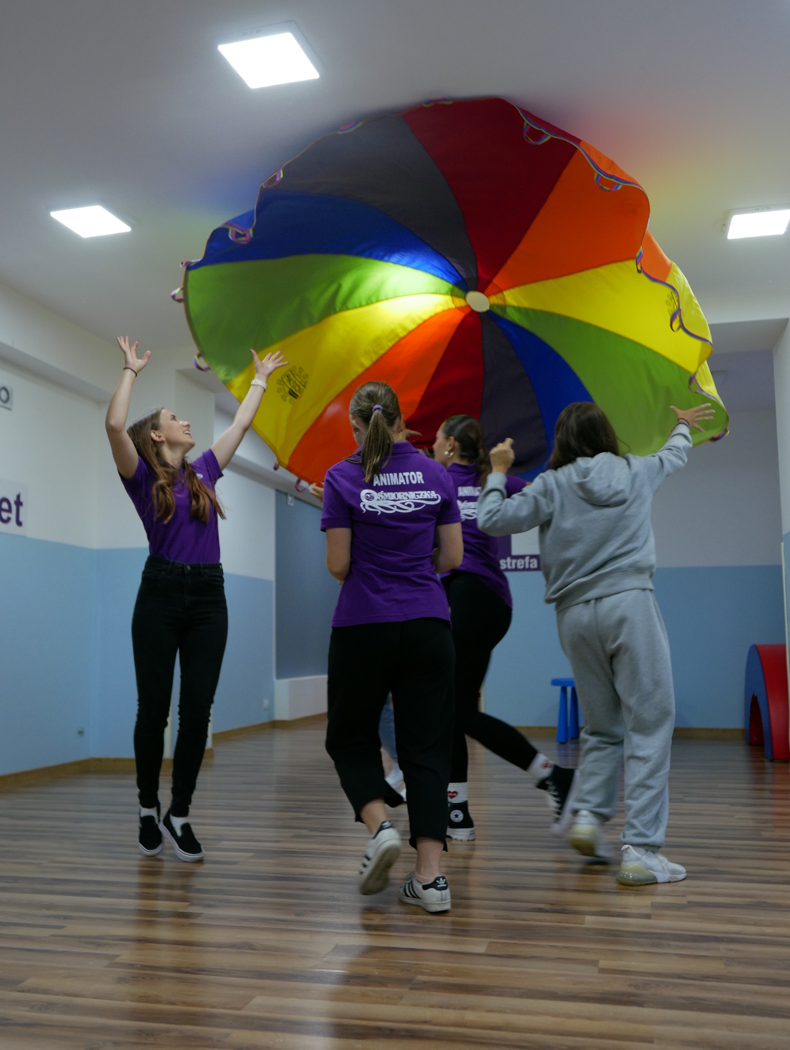 Grupa osób w fioletowych koszulkach unosi nad głowami dużą, kolorową chustę animacyjną w jasnym pomieszczeniu z drewnianą podłogą.