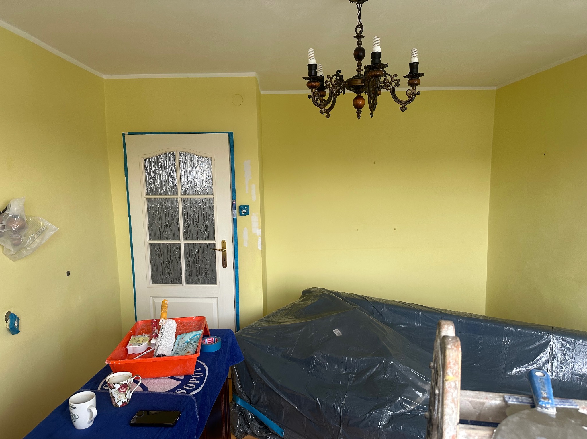 Pokój z żółtymi ścianami w trakcie przygotowania do malowania: meble przykryte folią, taśma malarska zabezpiecza framugę drzwi, widoczny kuweta z wałkami i pędzlami, ślady po szpachlowaniu na ścianie.