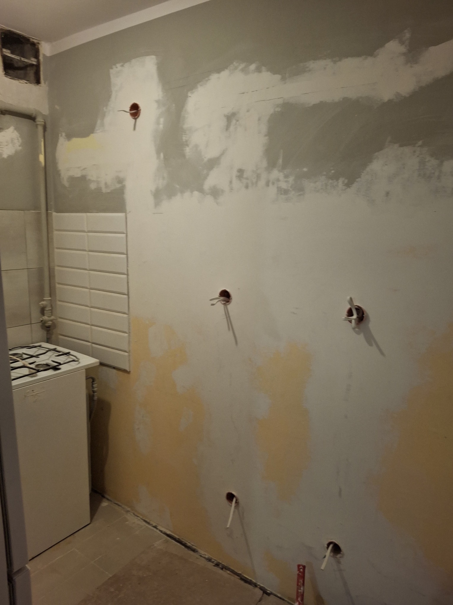 Przygotowanie ściany do malowania w kuchni: częściowo zaszpachlowana ściana z widocznymi instalacjami elektrycznymi i gazowymi, fragment kuchenki i płytek.