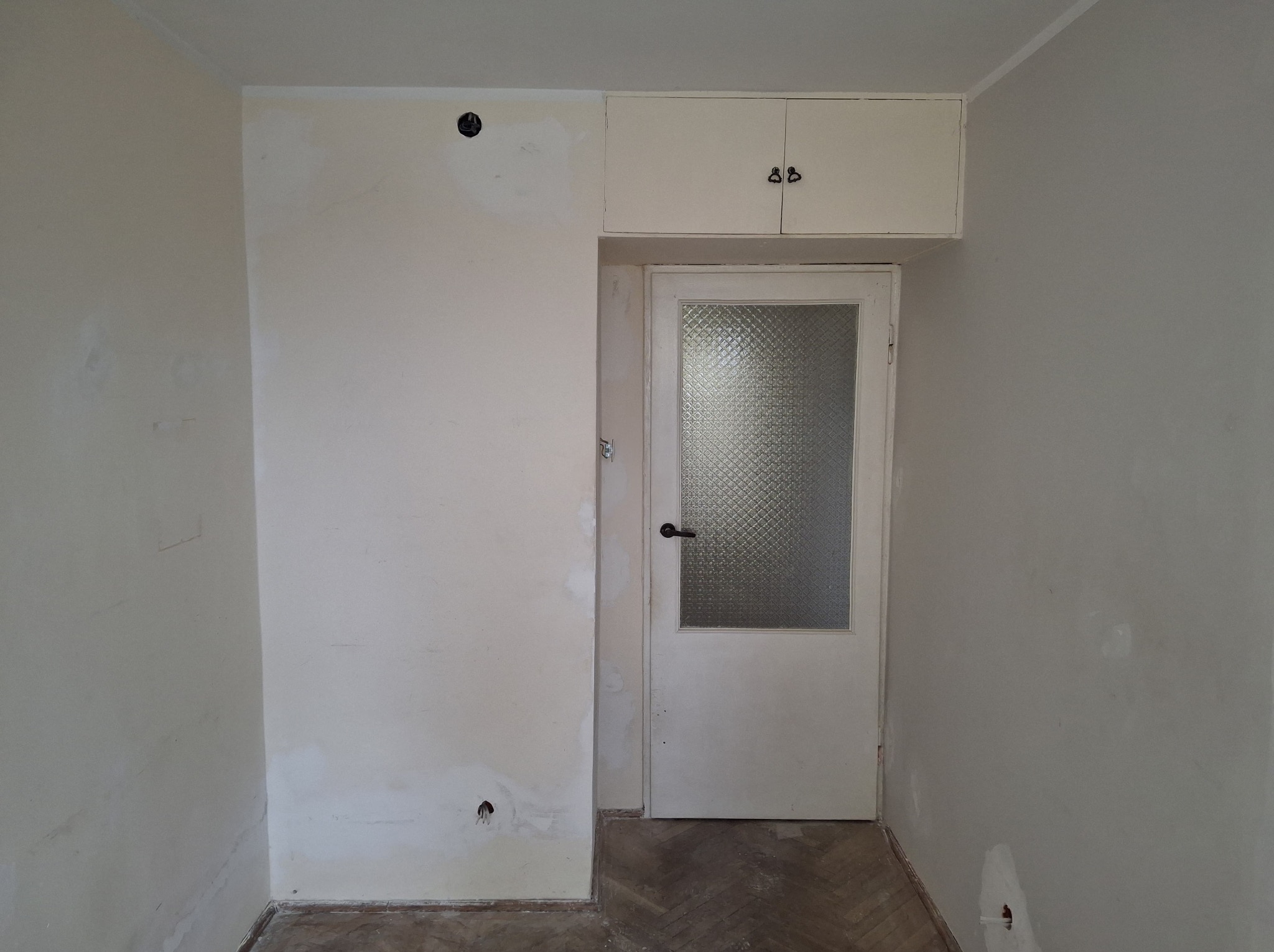 Pokój w trakcie przygotowania do malowania: ściany z widocznymi śladami po szpachlowaniu, fragment podłogi z parkietem, białe drzwi z szybą ornamentową i szafka nad nimi.