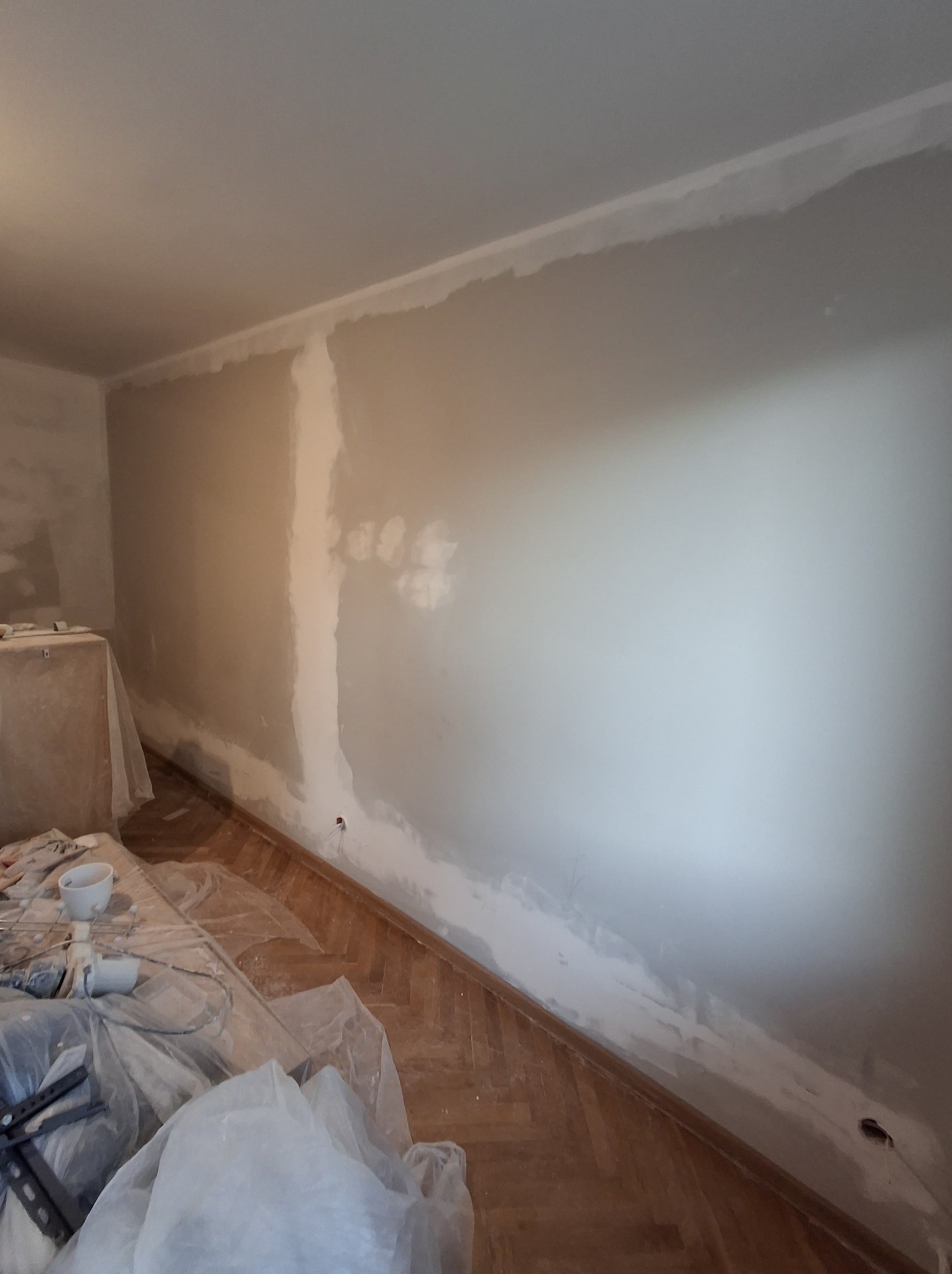 Pokój w trakcie remontu: częściowo wyszpachlowana ściana z widocznymi niedoskonałościami, zabezpieczona podłoga z parkietu i narzędzia malarskie.