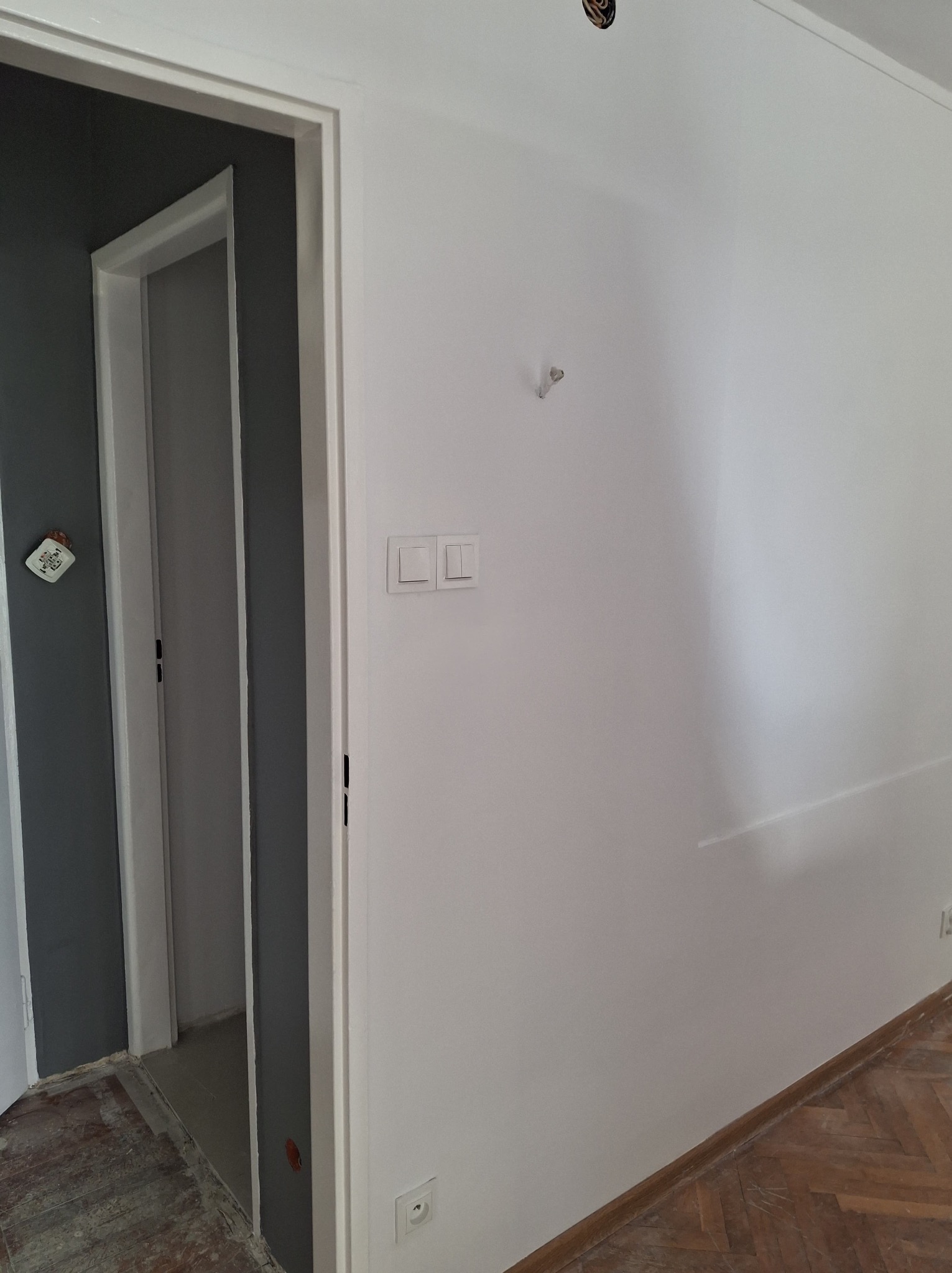 Biała ściana po odświeżeniu z widocznymi włącznikami światła i gniazdkiem elektrycznym, fragment parkietu przy podłodze oraz drzwi do pomalowanego na szaro pomieszczenia obok.