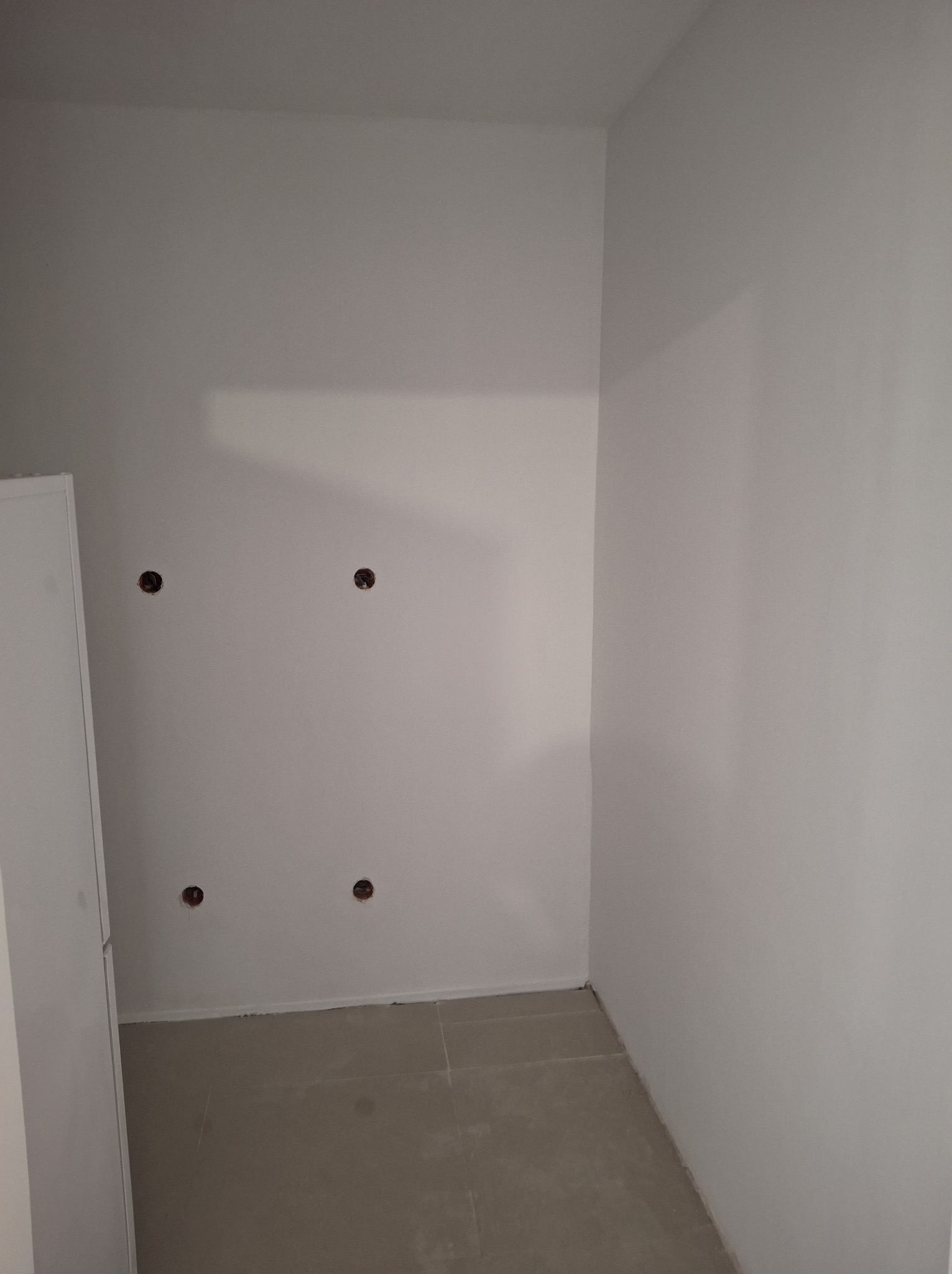 Pomieszczenie po malowaniu na biało, widoczne cztery otwory w ścianie, prawdopodobnie po gniazdkach elektrycznych, oraz fragment białej szafki.