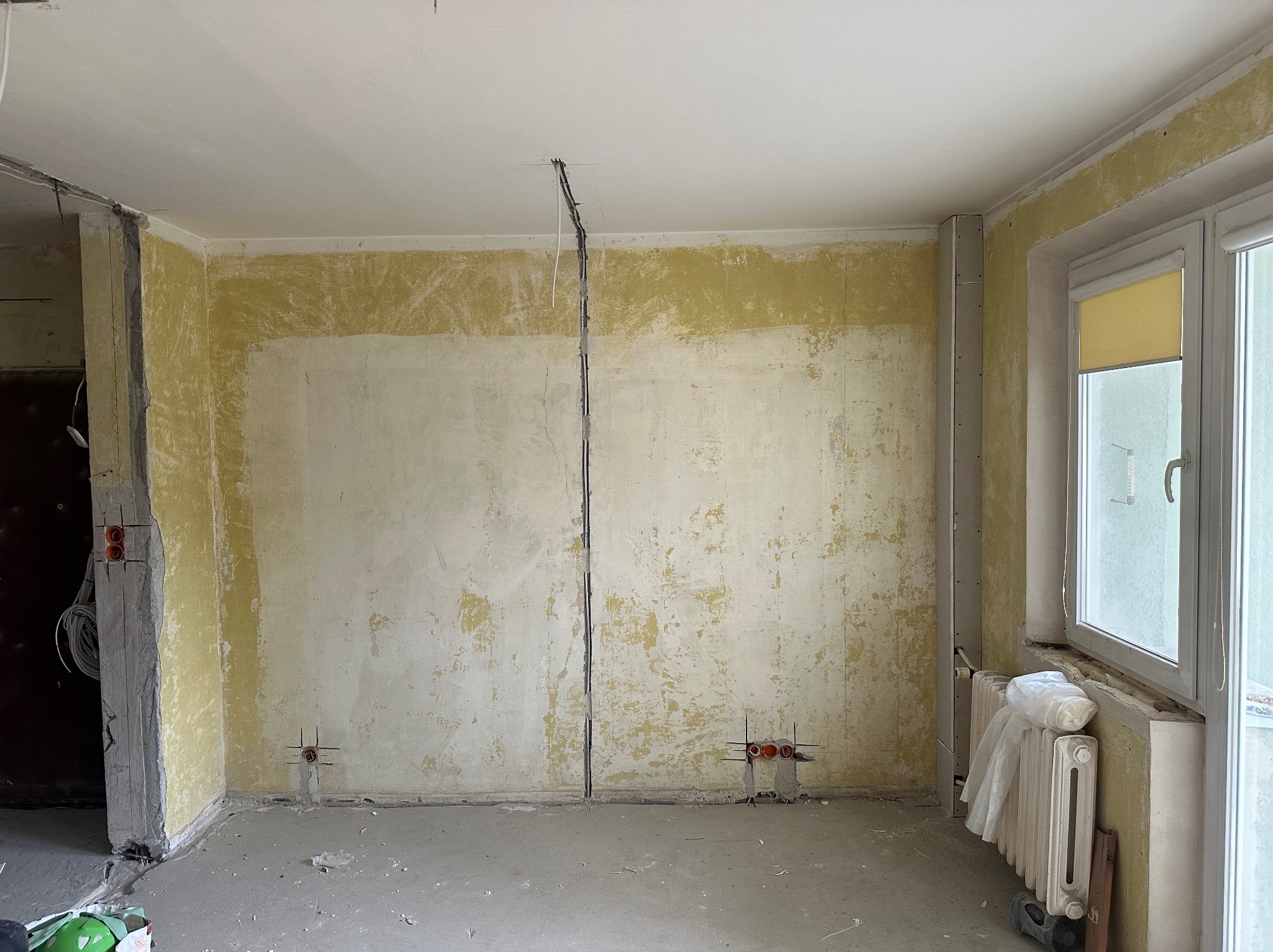 Pokój w trakcie remontu: ściany przygotowane do szpachlowania, widoczne instalacje elektryczne i grzewcze. Okno z roletą. Elementy osprzętu elektrycznego.