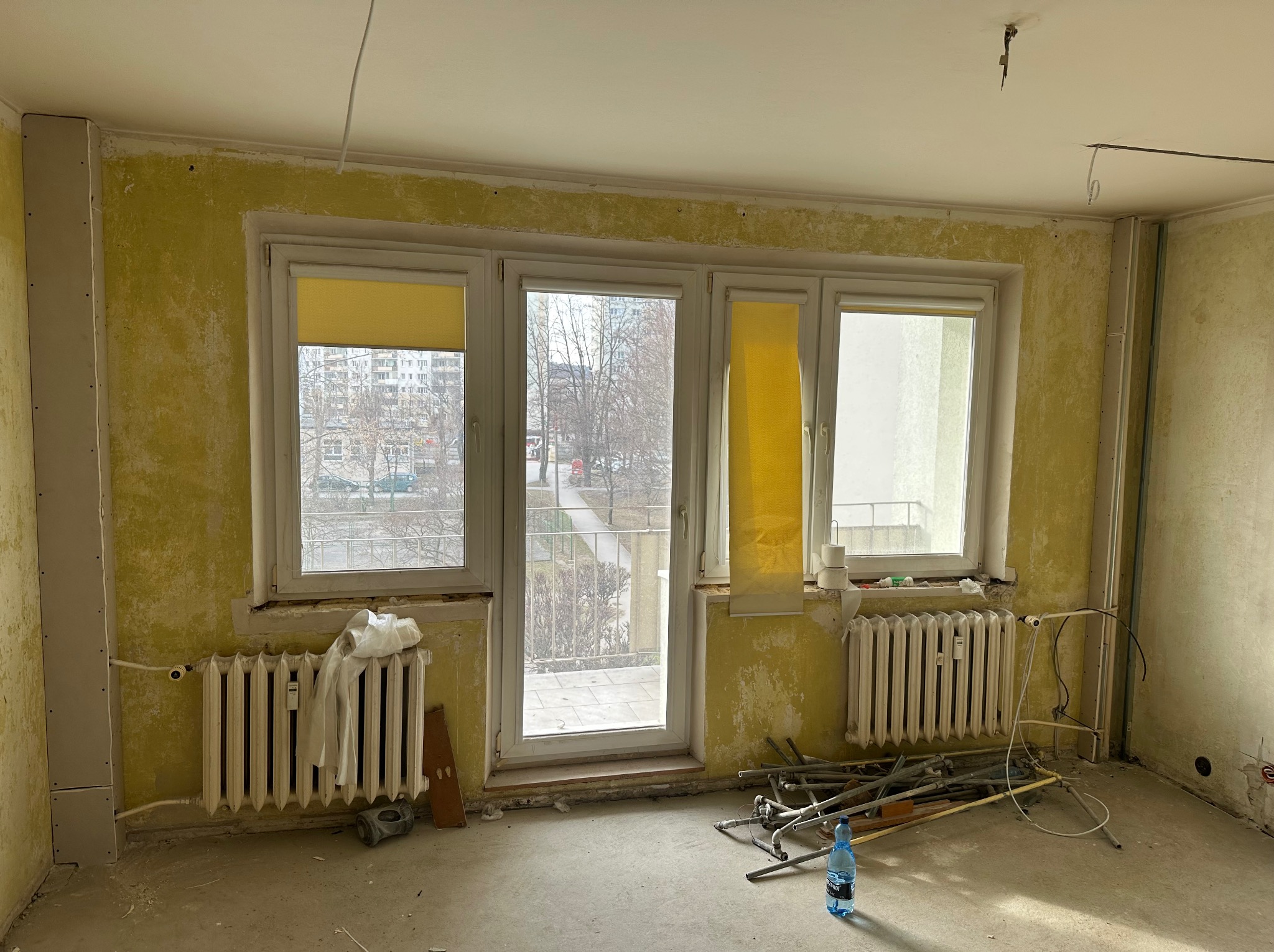 Pokój w trakcie remontu: ściany z resztkami farby, okna z roletami, grzejniki i rury na podłodze. Widoczne przygotowania do prac wykończeniowych.
