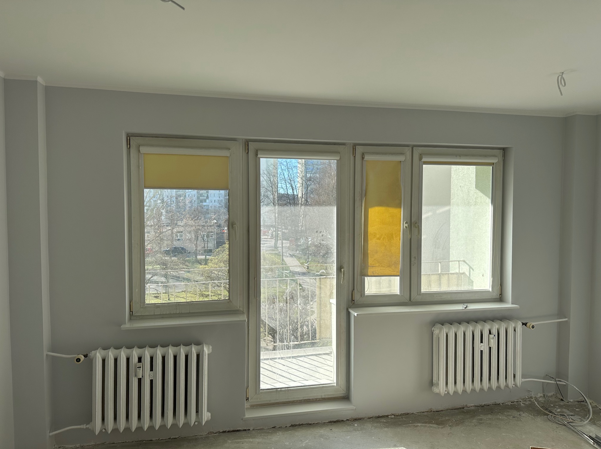 Jasne, pomalowane na szaro wnętrze z trzema oknami, grzejnikami i żółtymi roletami. Widoczne niedociągnięcia na podłodze sugerują prace wykończeniowe.