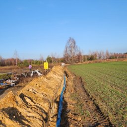 Nowoczesne technologie oze - Wykop w ziemi z niebieską rurą na dnie, widoczny fragment pola i las w tle, osoba w oddali.