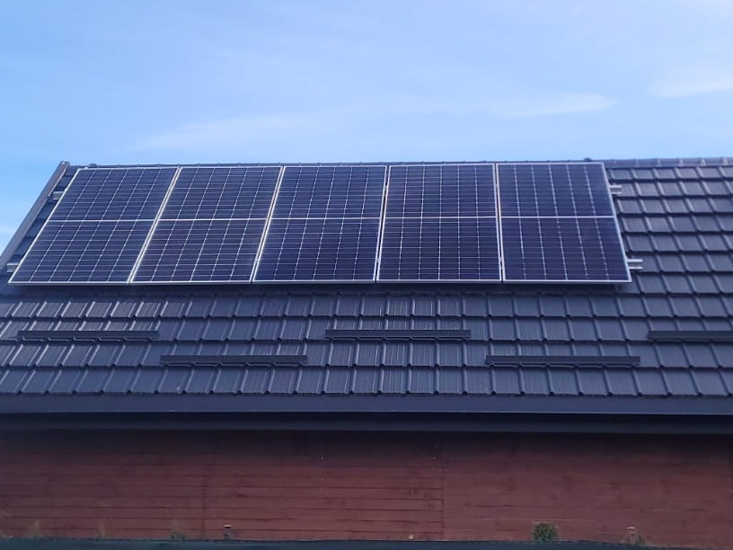 Panele fotowoltaiczne zamontowane na dachu z ciemnej dachówki, widoczne spod lekkiego kąta, na tle błękitnego nieba.