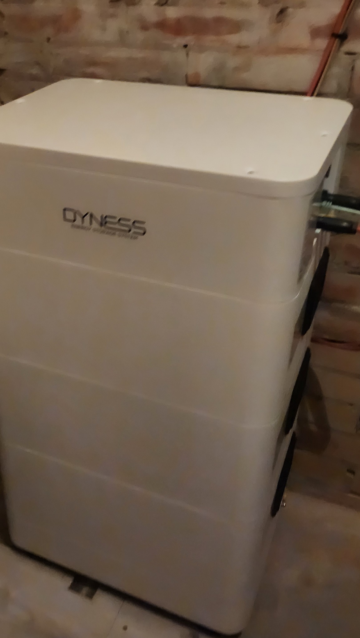 Biały akumulator energii Dyness, widok z boku, zainstalowany w pomieszczeniu z ceglaną ścianą w tle.