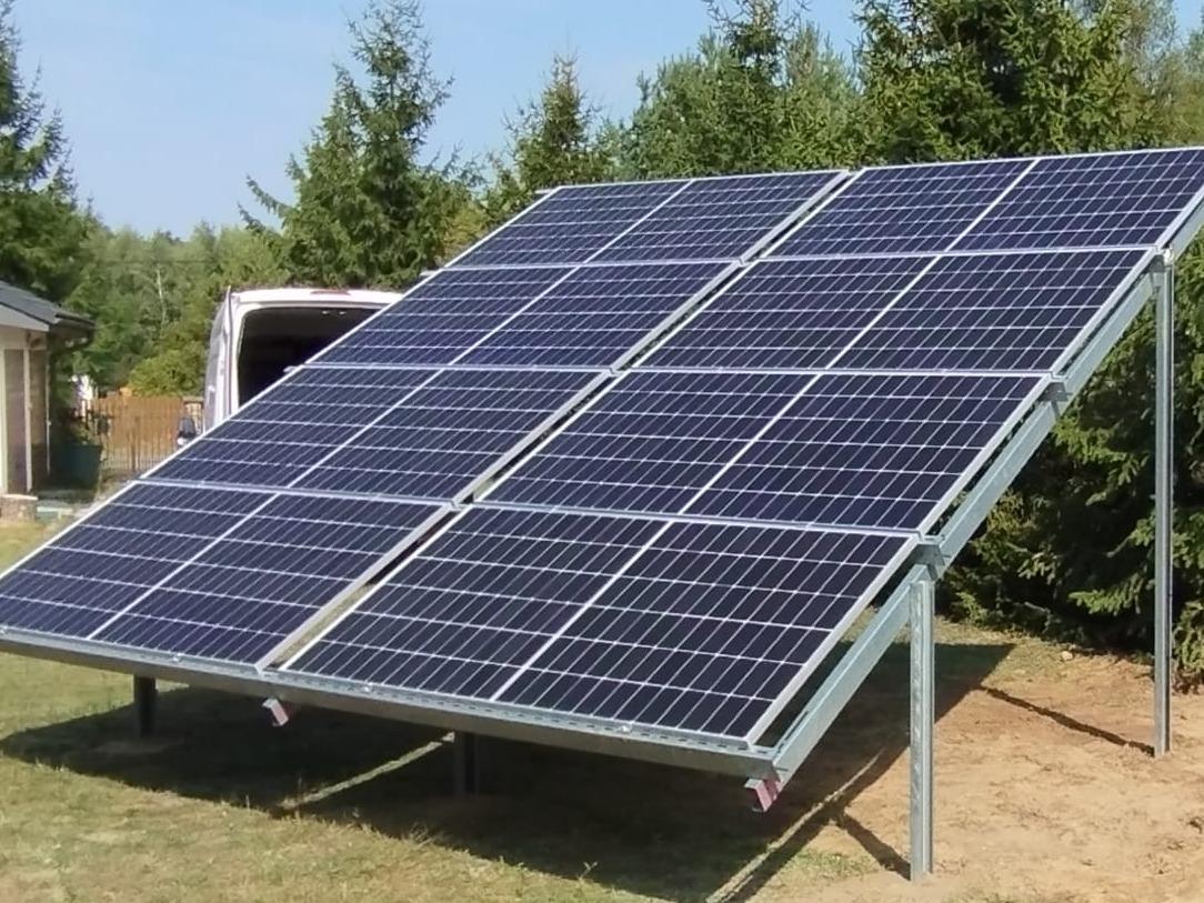 Panel fotowoltaiczny ustawiony na metalowej konstrukcji na trawniku przed domem, widoczny biały van w tle.