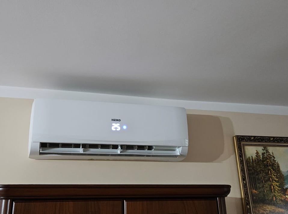 Biały klimatyzator marki Heiko zamontowany na ścianie nad ciemnobrązową komodą, z widocznym obrazem w złotej ramie po prawej stronie.