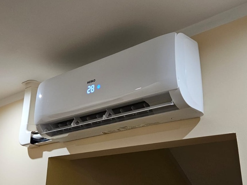 Biały klimatyzator marki HEIKO z wyświetlaczem temperatury 28 stopni Celsjusza zamontowany na beżowej ścianie, widoczny fragment szafki poniżej.