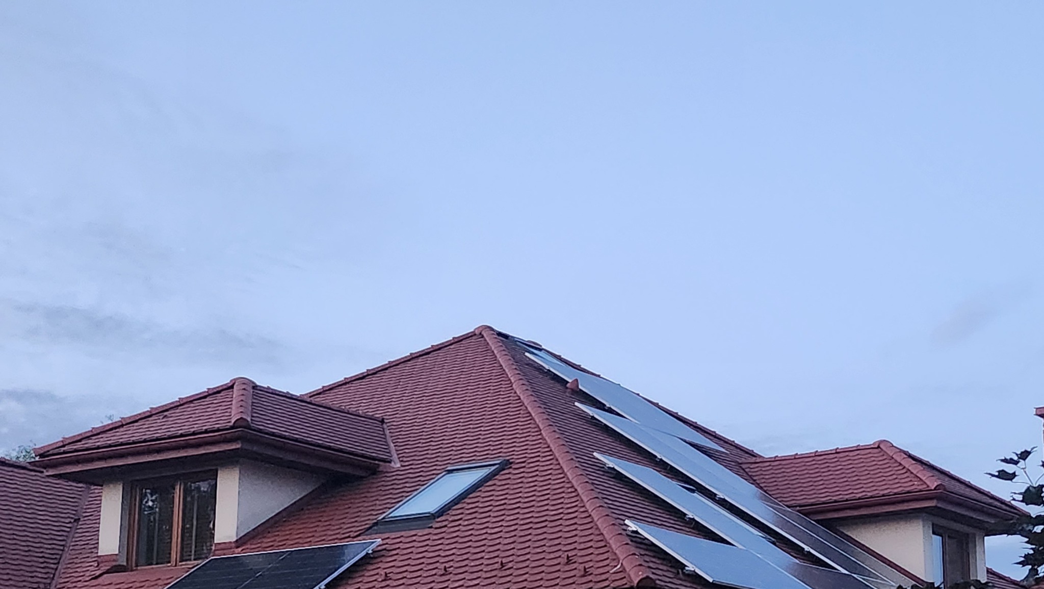 Czerwony dach domu z dachówką ceramiczną i zamontowanymi panelami fotowoltaicznymi, widoczne okno dachowe i fragment elewacji, pochmurne niebo w tle.