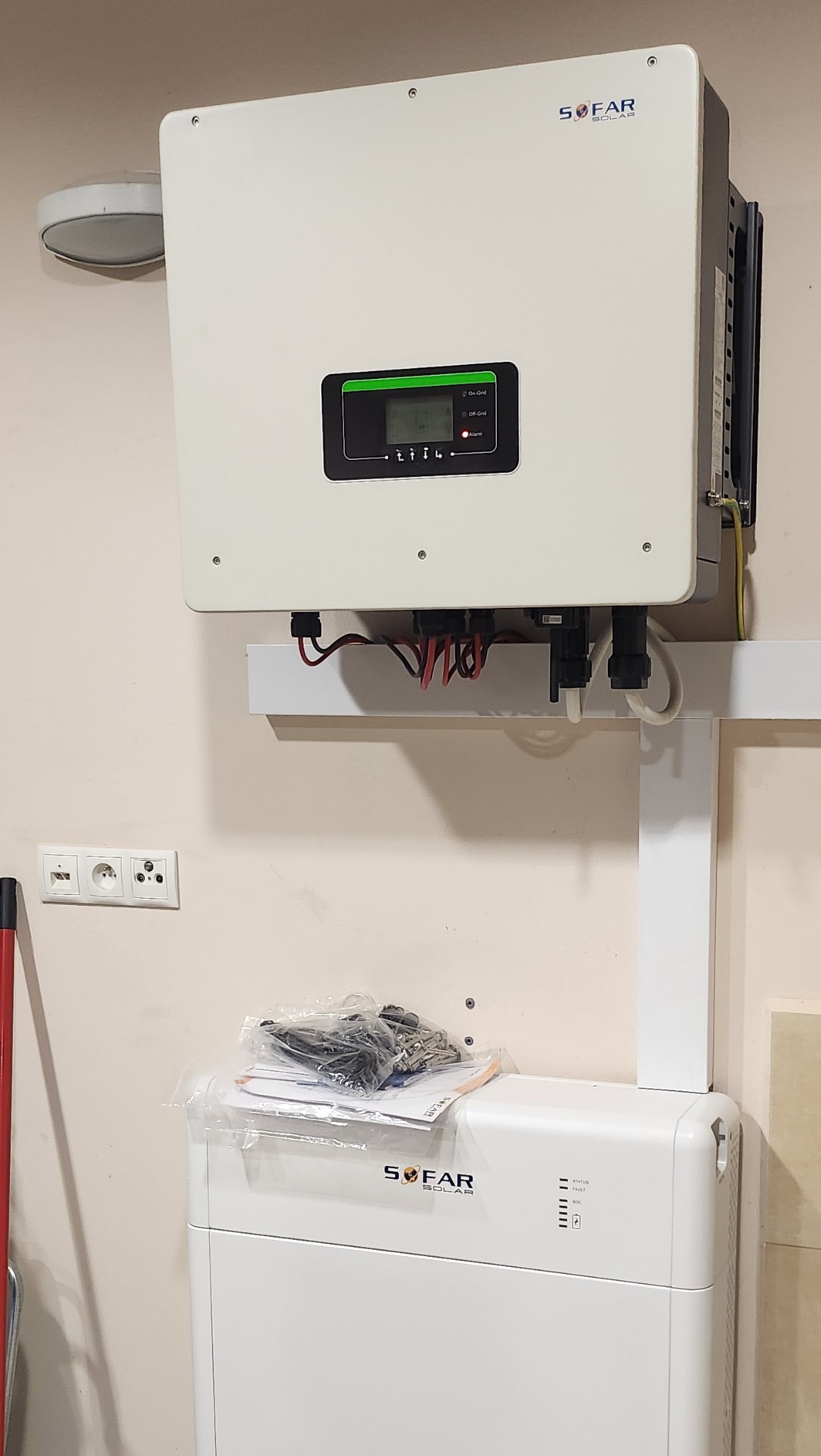 Biały inwerter fotowoltaiczny marki Sofar Solar zamontowany na ścianie, z widocznymi przewodami, obok gniazdka elektrycznego i akumulatora z instrukcją w folii na górze.