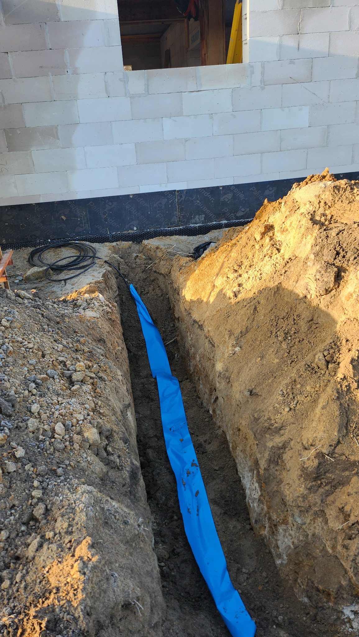 Wykop z niebieską folią ochronną ułożoną wzdłuż fundamentu budynku z betonu komórkowego, widoczne kable. Etap prac ziemnych przed instalacją.