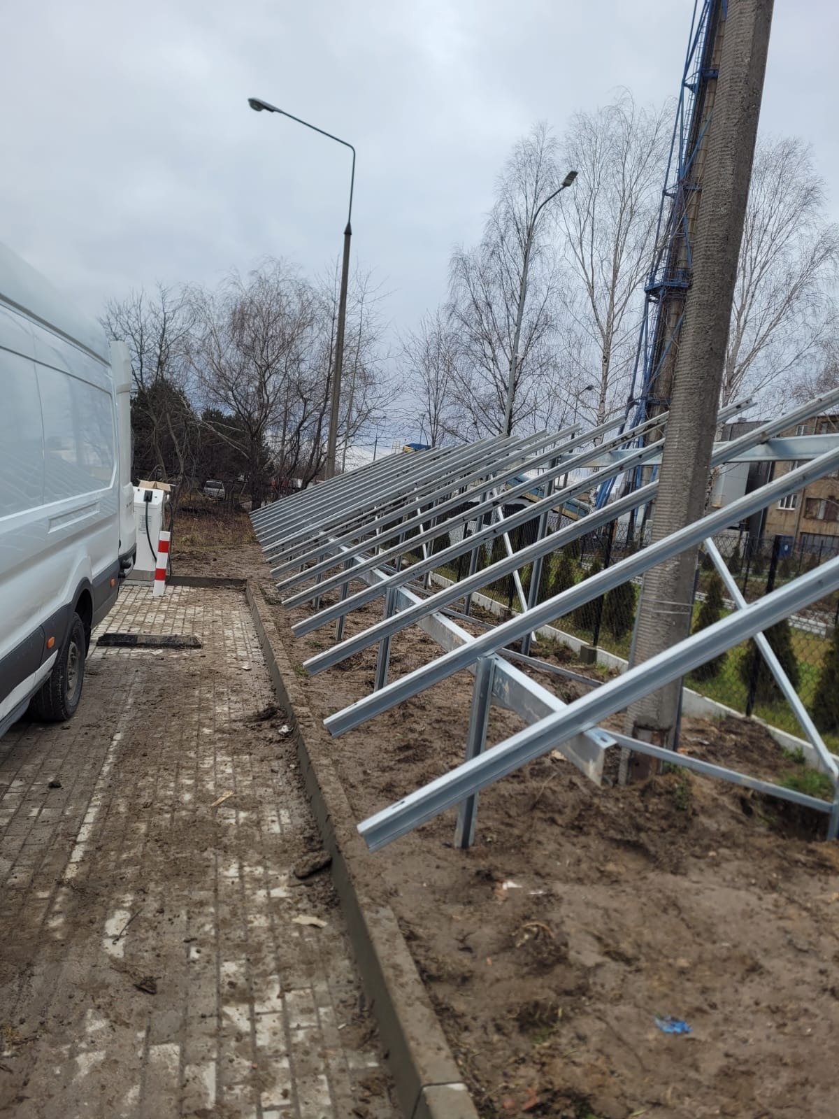 Stalowa konstrukcja wsporcza pod panele fotowoltaiczne, widoczna z perspektywy ziemi, z zaparkowanym białym vanem w tle i słupem energetycznym po prawej stronie. Instalacja w trakcie montażu...