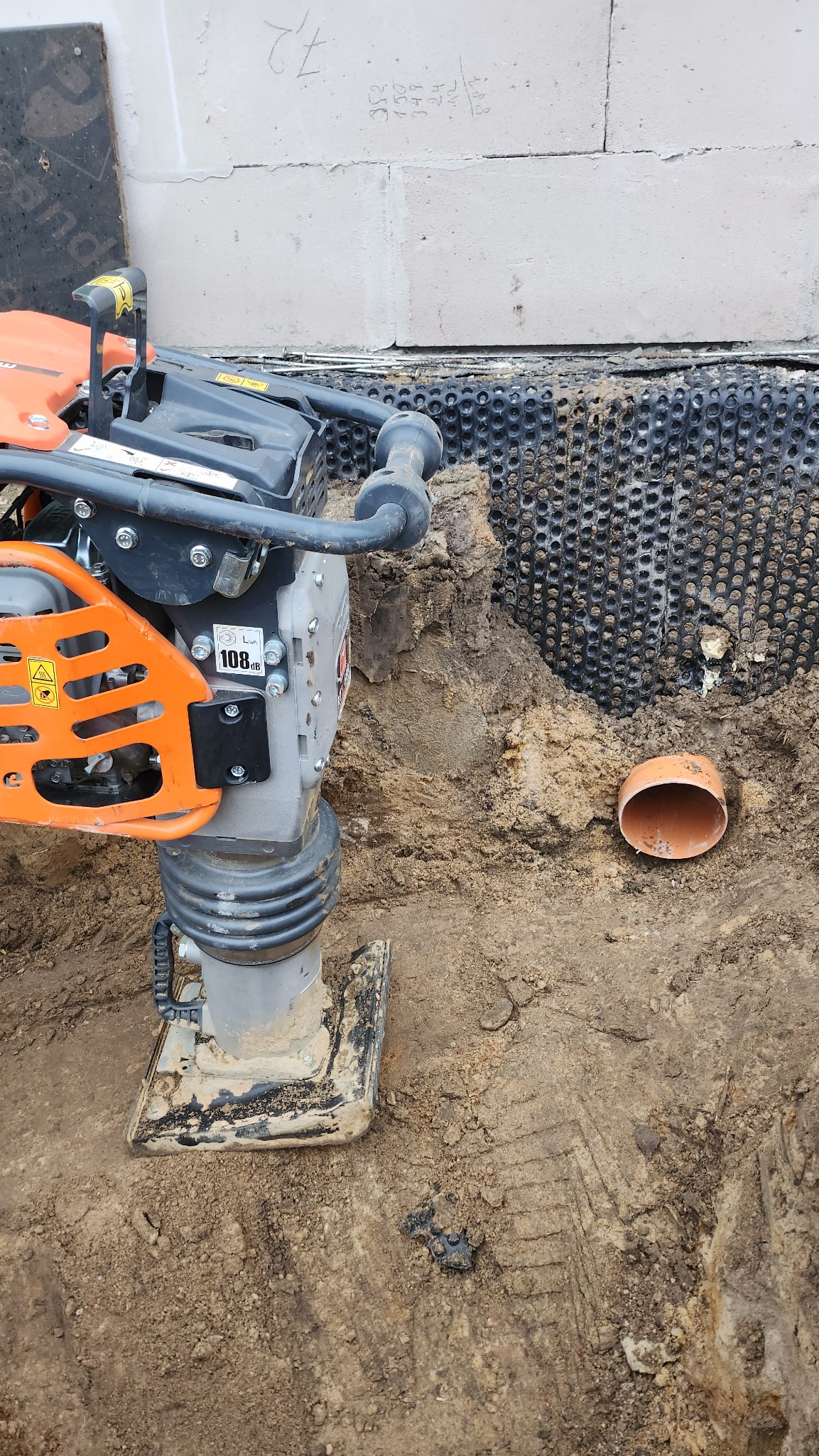 Pomarańczowo-szara ubijarka mechaniczna do gruntu przy fundamentach budynku z izolacją pionową i fragmentem rury kanalizacyjnej w wykopie.