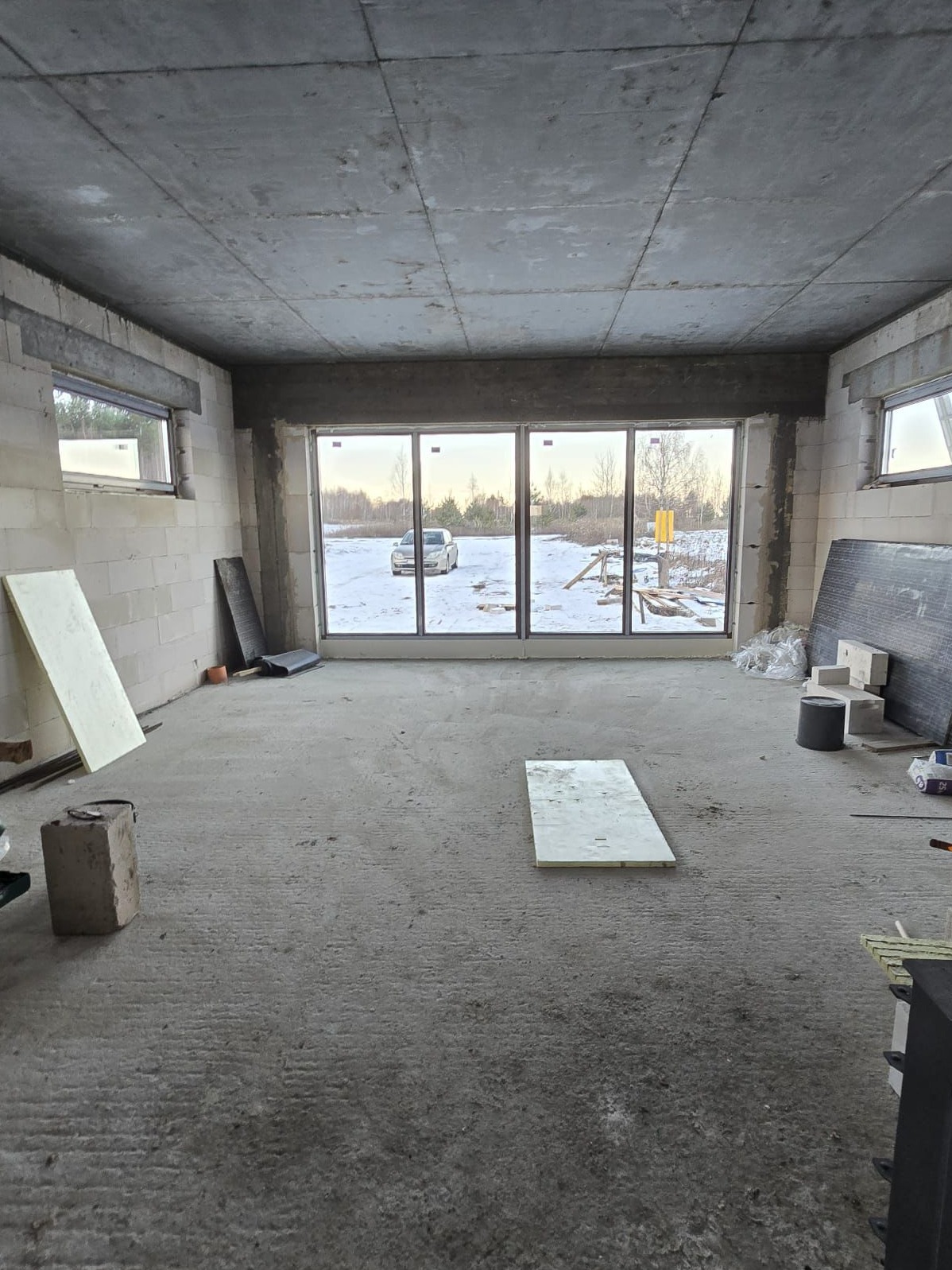 Surowy stan wnętrza domu w trakcie budowy, widoczne bloczki, wylewka, okna z widokiem na zimowy krajobraz, fragmenty izolacji i materiałów budowlanych.