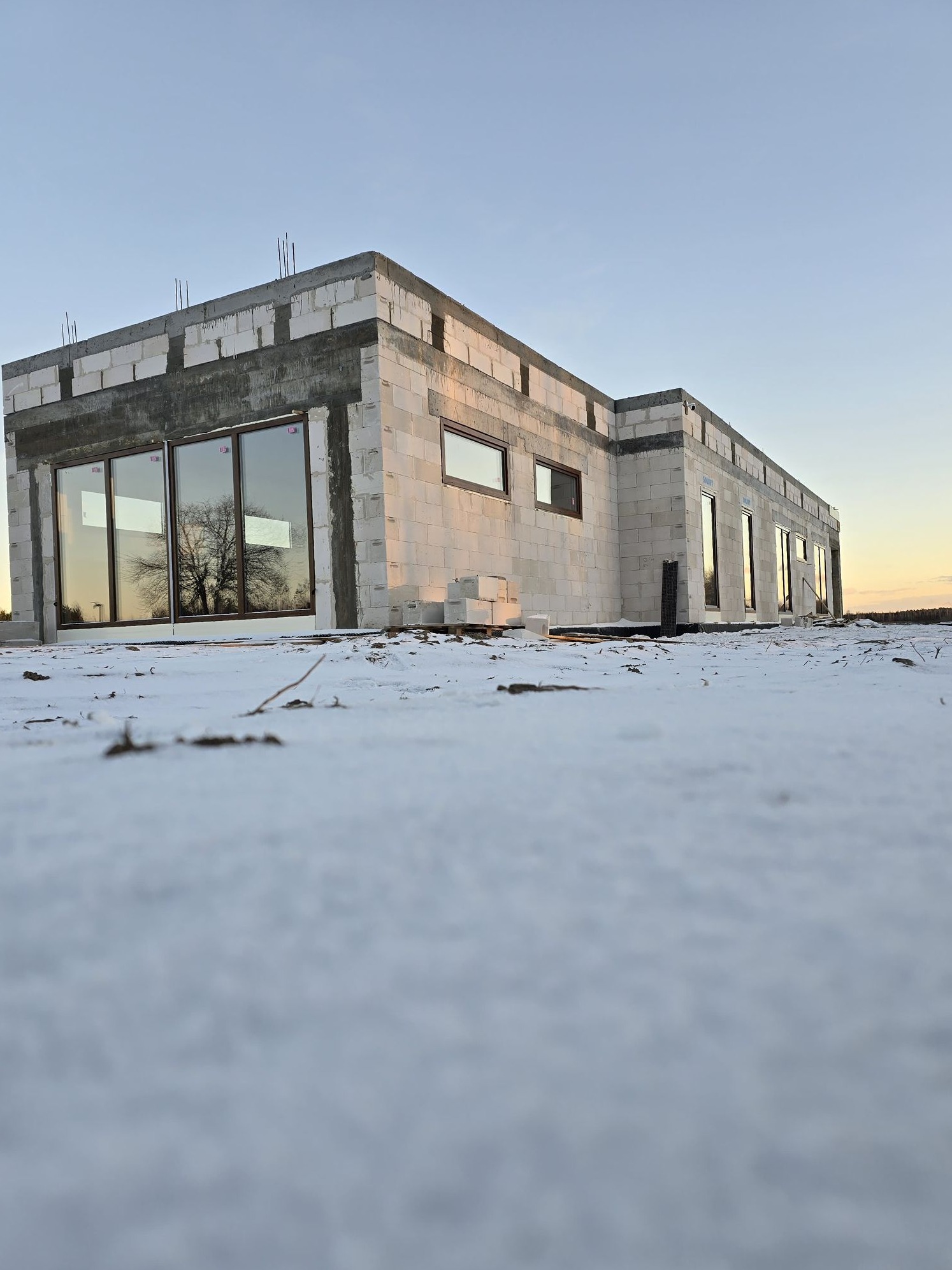 Surowy stan domu z białych bloczków w zimowej scenerii, widoczne duże okna z brązowymi ramami odbijające drzewo, wystające pręty zbrojeniowe.