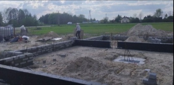 Budowa fundamentów domu: widoczne bloczki fundamentowe, izolacja przeciwwilgociowa, zbrojenie, piasek oraz pracownik w oddali.