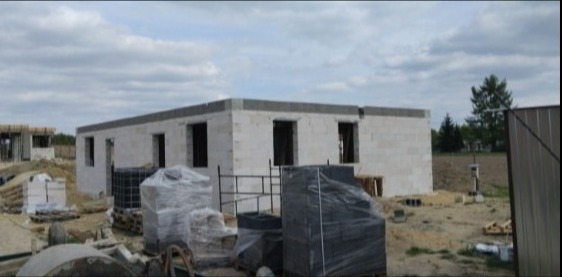 Budowa domu jednorodzinnego w stanie surowym otwartym z białych bloczków, widoczne materiały budowlane i palety, pochmurne niebo.