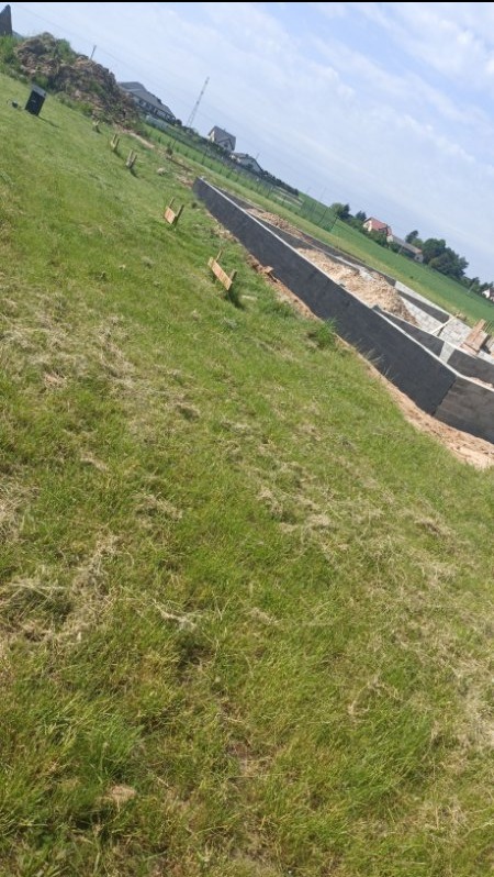 Wylane fundamenty pod budowę domu na zalesionym terenie, widoczne drewniane stemple, w tle sterta ziemi i zabudowania.