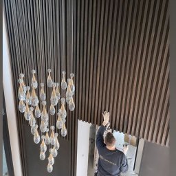 Dom&Greg - Montaż paneli ściennych z ciemnego drewna w wysokim pomieszczeniu, widoczny pracownik w rękawicach na tle jasnych ścian i wiszący żyrandol z kryształkami