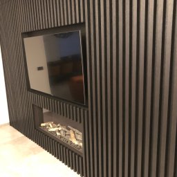 Dom&Greg - Ściana z czarnych, pionowych lameli z wnęką na telewizor i imitacją kominka z dekoracyjnymi polanami.