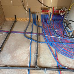 Dom&Greg - Instalacja ogrzewania podłogowego w trakcie realizacji: liczne czerwono-niebieskie rury prowadzone do rozdzielaczy na ścianie, czarne rury kanalizacyjne przy podłodze, surowa ściana z cegły i płyty...