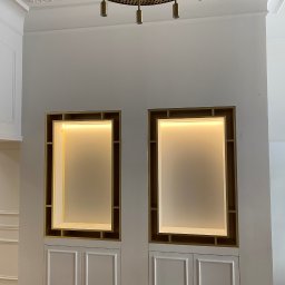 Dom&Greg - Dwa podświetlane wnęki z półkami i szafkami w dolnej części, umieszczone w białej ścianie z dekoracyjną listwą sufitową i złotym żyrandolem.