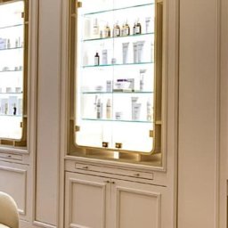 Dom&Greg - Ściana z wbudowanymi, podświetlanymi witrynami z kosmetykami i kremami w eleganckim salonie kosmetycznym, widoczny fragment szklanego stolika i parkietu.