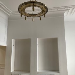 Dom&Greg - Wnętrze z białymi ścianami, sztukaterią sufitową i wnękami z szafkami, oświetlone przez okrągły, złoty żyrandol z punktowymi otworami.